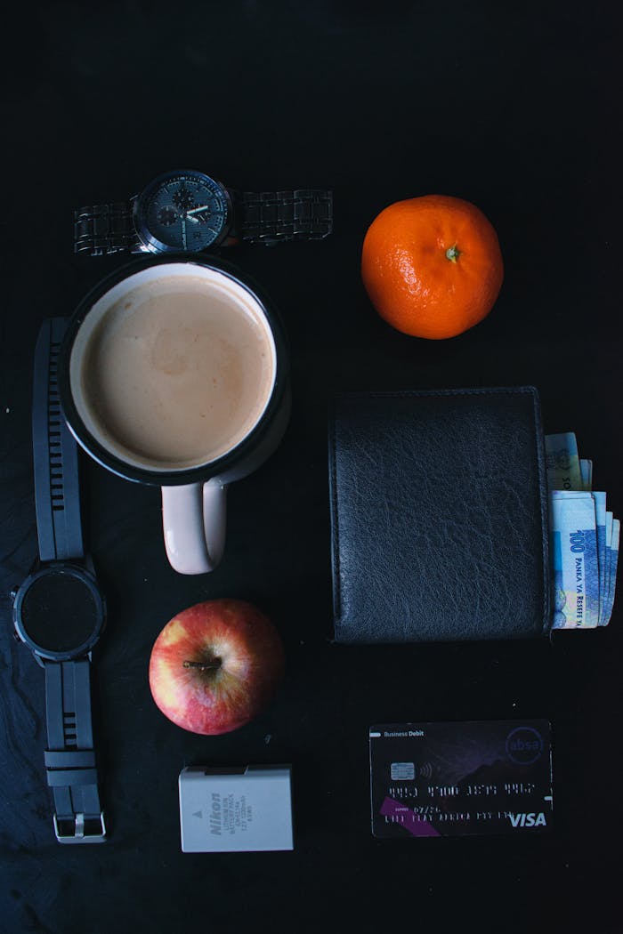 Overhead view of daily essentials like watch, wallet, and fruit on dark surface.